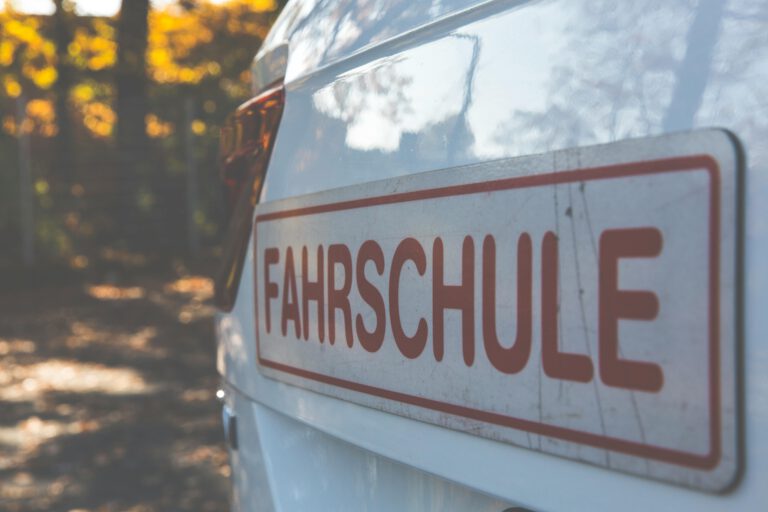 A side view of a white car with a Fahrschule sign, outdoors with autumn foliage.